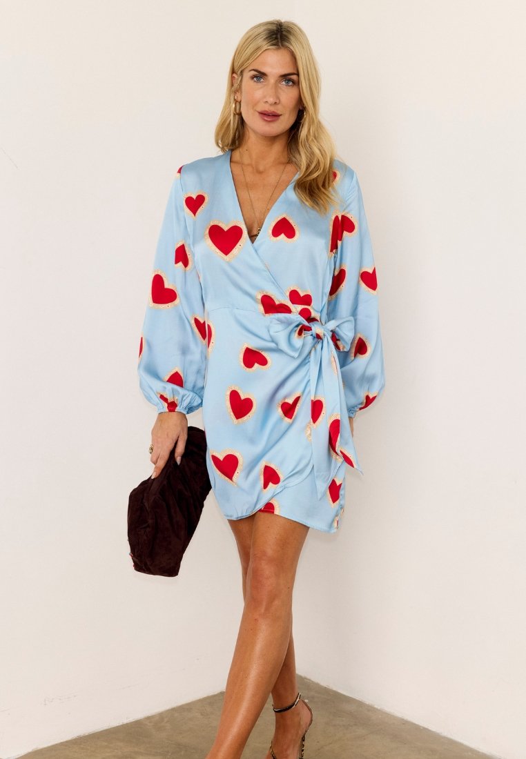 Γυναίκα που στέκεται μπροστά σε μπεζ τοίχο φορώντας ένα μπλε φόρεμα τύπου κρουαζέ με κόκκινες καρδιές, κρατώντας μια σκούρα καφέ τσάντα χειρός στο αριστερό της χέρι.