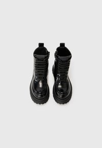 Bottes militaires en cuir noir avec une finition brillante, design à lacets, cols rembourrés et semelles en caoutchouc épaisses et texturées. Fermeture éclair sur le côté pour un enfilage facile.