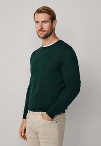 Maglione verde a girocollo realizzato in un morbido tessuto, con maniche lunghe e un colletto interno bianco a contrasto. Abbinato a pantaloni chiari.