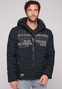 Giubbotto piumino nero con chiusura a zip frontale, cappuccio e design a trapunta orizzontale, con testo grafico sul petto e logo sulla manica.