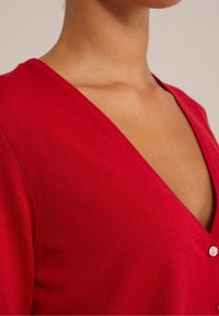 Röd cardigan med djup V-ringning, mjukt stickat material och dekorativ knappdetalj vid stängningen. Slätt tyg och figursydd design.