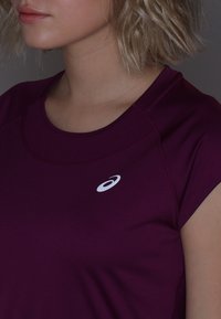 Mörkröd träningstopp med korta ärmar, med rund halsringning och en liten vit logotyp på vänster bröstområde. Slät tygtextur.