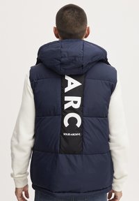 Navy plavi puffer prsluk s kapuljačom, s crnom panelom na leđima na kojem je ispisano "ARC" i "SOLID ARCHIVE" u bijeloj boji. Teksturan i podstavljen.