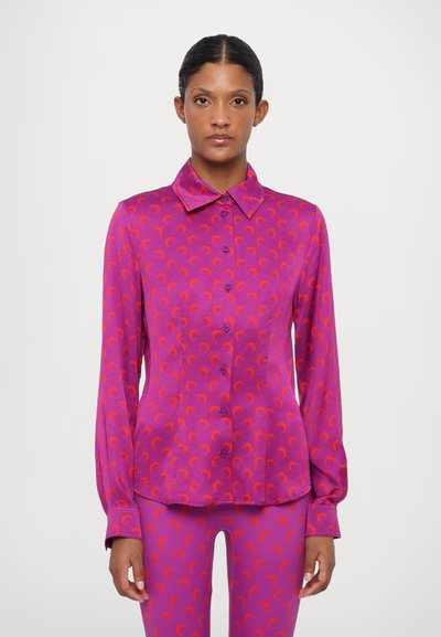 Chemisier fuchsia à manches longues, avec un boutonnière sur le devant, et un motif de croissant de lune répétitif en orange. Fabriqué en tissu lisse et brillant.