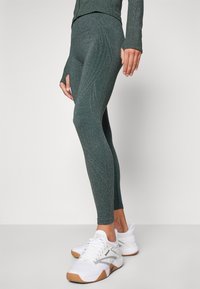 Leggings vert texturé avec une subtile brillance, dotés d'une taille haute et d'une coupe ajustée. Associés à des chaussures de sport blanches et des accents gris.
