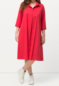 Punainen, polvenpituinen mekko, jossa on kaulus ja kolmanneksen pituiset hihat, valmistettu sileästä kankaasta. Yhdistetty valkoisiin korkeisiin tennareihin.