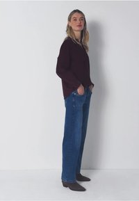 Viininpunainen pitkähiha toppi rentolla leikkuulla, yhdistettynä sinisiin leveälahkeisiin farkkushortseihin. Ruskeat nilkkurit täydentävät asua. Yksinkertainen valkoinen tausta.