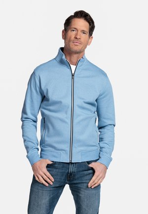 Man die een lichtblauwe jas met ritssluiting en zakken draagt en een blauwe spijkerbroek, staand met handen bij de zakken tegen een witte achtergrond.