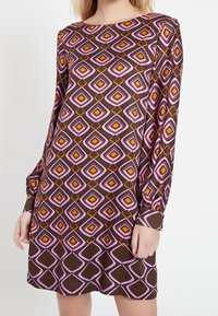 Modell bär en långärmad klänning med ett geometriskt mönster i brunt, rosa och orange, stående mot en vit bakgrund.