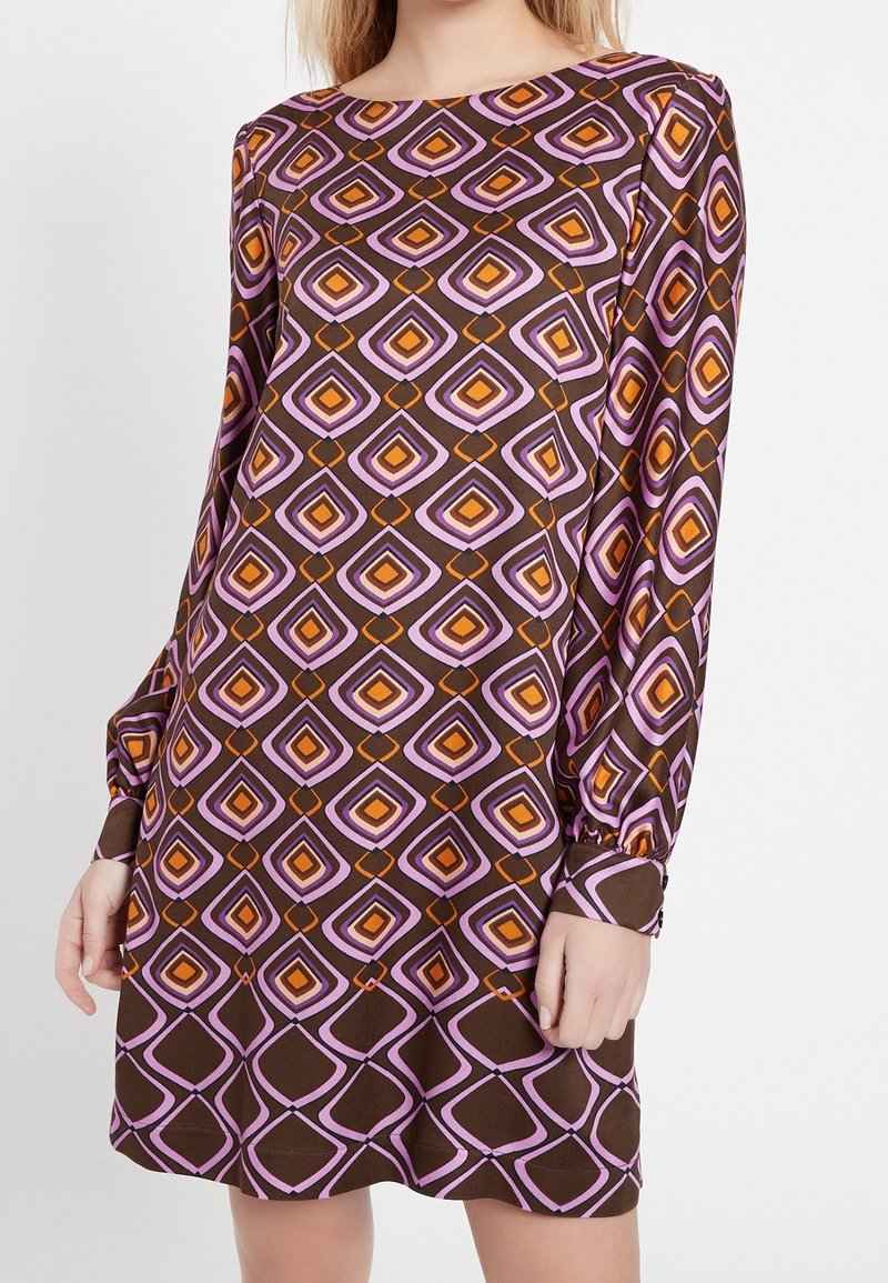 Modell bär en långärmad klänning med ett geometriskt mönster i brunt, rosa och orange, stående mot en vit bakgrund.