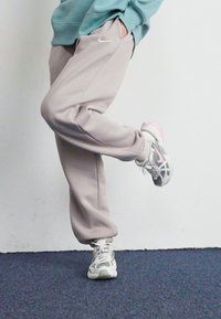 En person iført beige sweatpants og hvide sneakers med lyserøde detaljer løfter det ene ben mod en ensfarvet, lys væg og et mørkt tæppe.