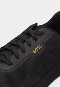 Nahaufnahme eines schwarzen Sneakers mit strukturiertem Stoff, schwarzen Schnürsenkeln und goldenem "BOSS"-Logo an der Seite in der Nähe der Schnürung.