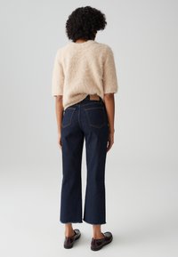 Lichtbeige fluffy trui met korte mouwen, gecombineerd met donkerblauwe cropped jeans met een ruwe zoom en achterzakken. Bruine platte schoenen.