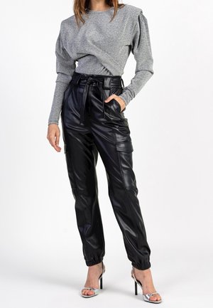 Femme portant un haut gris à manches longues et un pantalon cargo noir en similicuir taille haute avec poignets élastiques et sandales argentées à talons hauts.