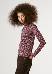 Langarmtop van donkere stof met een roze bloemenpatroon. Heeft een hoge hals en een getailleerd ontwerp, gecombineerd met donkerbruine broek.