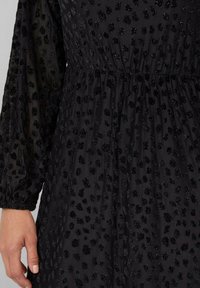 Zbliżenie czarnej sukienki z przezroczystymi długimi rękawami i subtelnym wzorem w panterkę, z aksamitnymi plamkami o teksturze.