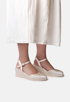 Feet wearing satin beige wedge espadrille sandals with ankle straps, shown with a white textured midi dress.