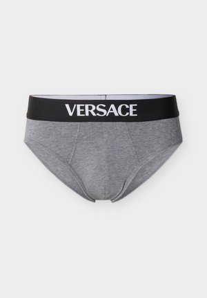 Skjorteløs mann iført grå Versace-boxershorts med svart midjebånd, stående foran en ensfarget lys grå bakgrunn.