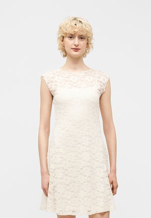 Femme blonde aux cheveux courts et bouclés portant une robe blanche sans manches en dentelle à motif floral, debout devant un fond clair et uni.