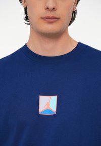 Tumši zila kokvilnas T-kreklu ar kvadrātveida logo, kurā redzams lēkājošs tēls oranžās un zilās akcenta krāsās, ar apaļu kakla izgriezumu un īsām piedurknēm.