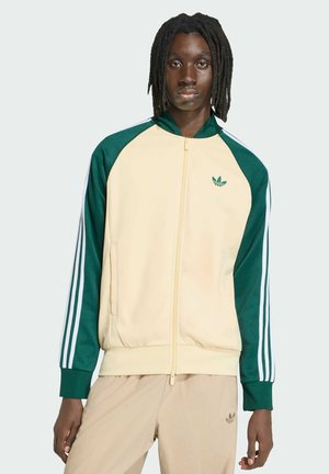 Młoda osoba z średniej długości dredami, ubrana w kremowo-zieloną kurtkę dresową Adidas oraz beżowe spodnie Adidas, stojąca na jednolitym tle.
