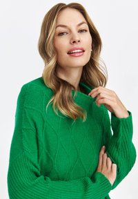 Kobieta z falującymi blond włosami w jasnej zielonej swetrze w warkocze, trzymająca kołnierz i pewnie patrząca w obiektyw.