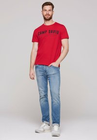 Red short-sleeve t-shirt with "CAMP DAVID" graphic in black, paired with light blue denim jeans and white shoes.