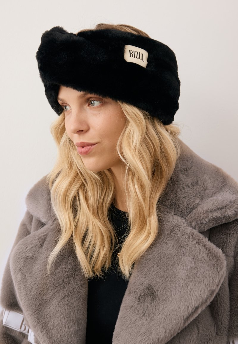 Czarna opaska na głowę z sztucznego futra o fakturowanej powierzchni i prostokątnym beżowym metku z napisem "BIZUU". Nosi ją osoba w szarej futrzanej kurtce.