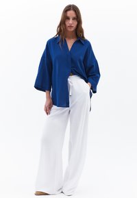 Camicia blu con bottoni, realizzata in tessuto testurizzato e maniche ampie, abbinata a pantaloni bianchi larghi in un materiale leggero.