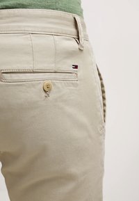 Beige puuvillahousut takaviistosta, joissa on yksinkertainen nappikiinnitys, takatasku, jossa on pieni logo-yksityiskohta, ja sileä pinta.