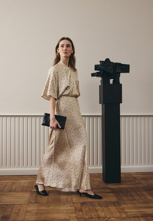 Nainen beigen pilkullisen mekon kanssa, joka pitelee mustaa kirjekuorilaukkua, seisoo minimalistisessa huoneessa modernin mustan veistoksen vieressä jalustalla.