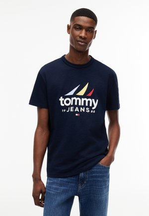 Młody mężczyzna ubrany w granatowy T-shirt Tommy Jeans z kolorowym logo żaglówki oraz niebieskie dżinsy, stoi z jedną ręką w kieszeni.