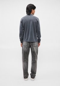 Γκρι μακρυμάνικο πουκάμισο με ομαλή υφή, συνδυασμένο με ξεθωριασμένα μαύρα τζιν και λευκά αθλητικά παπούτσια, προβαλλόμενο από πίσω.