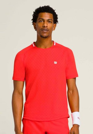 Mann trägt rotes kurzärmeliges Sportshirt und passende Shorts, mit weißem Handgelenksband, steht vor einem einfarbigen hellen Hintergrund.