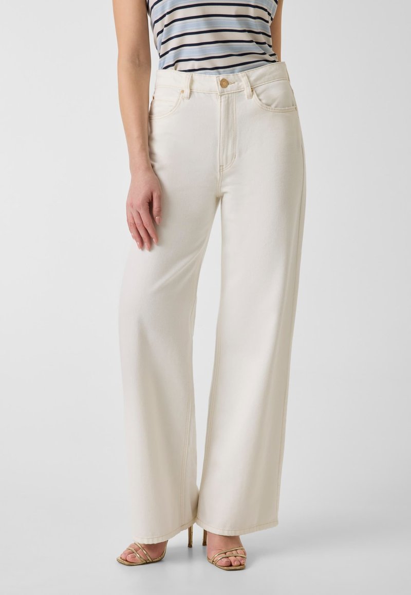 Femme portant un pantalon crème taille haute à jambes larges, un top rayé sans manches et des sandales dorées à talons hauts à brides, debout devant un fond uni.