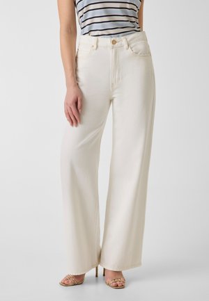 Femme portant un pantalon crème taille haute à jambes larges, un top rayé sans manches et des sandales dorées à talons hauts à brides, debout devant un fond uni.