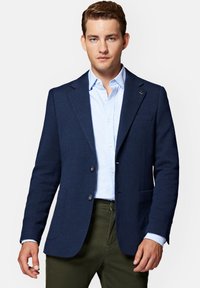 Granatowa, tkana marynarka z klapami, dwoma guzikami i fakturowanym materiałem. Nosi się ją na jasnoniebieskiej koszuli i oliwkowych spodniach.