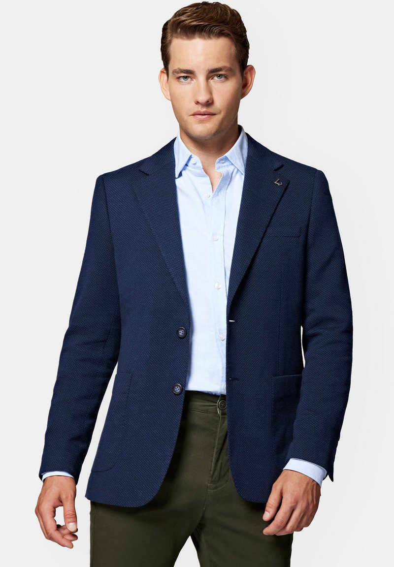 Granatowa, tkana marynarka z klapami, dwoma guzikami i fakturowanym materiałem. Nosi się ją na jasnoniebieskiej koszuli i oliwkowych spodniach.