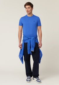 Blue cotton t-shirt with matching button-up shirt tied around the waist, paired with dark denim jeans and white sneaker accents.