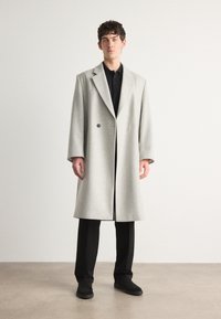 Palton din lână gri deschis, cu un design cu două rânduri de nasturi, cu rever ascuțite și două buzunare, purtat peste un polo negru și pantaloni.