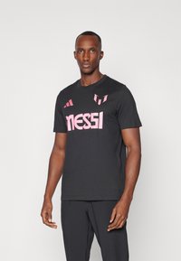 Camiseta negra de algodón con mangas cortas, presenta un llamativo texto en rosa "MESSI" y acentos del logotipo en el pecho. Diseño sencillo, sin patrones.