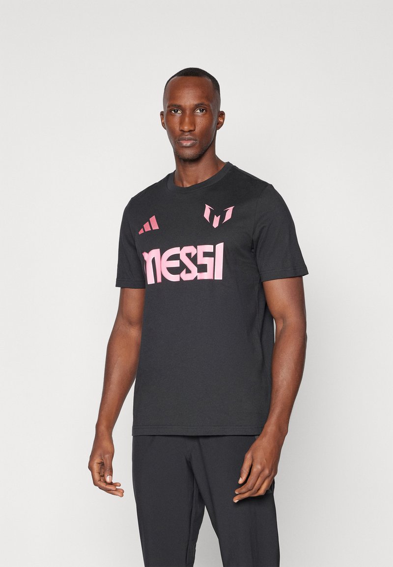 Camiseta negra de algodón con mangas cortas, presenta un llamativo texto en rosa "MESSI" y acentos del logotipo en el pecho. Diseño sencillo, sin patrones.