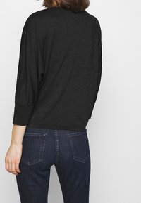 Kvinna som bär en glittrande svart långärmad tröja och mörkblå denimjeans, visad bakifrån mot en enkel vit bakgrund.