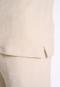Tessuto in spugna beige con una consistenza morbida, caratterizzato da un orlo leggermente allungato e uno spacco laterale per maggiore movimento e comfort.