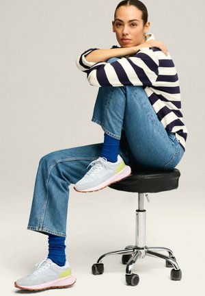 Zapatillas blancas con detalles en verde claro y suelas naranjas, combinadas con calcetines azules de canalé. Acompañadas de una blusa de rayas azul marino y jeans azules.