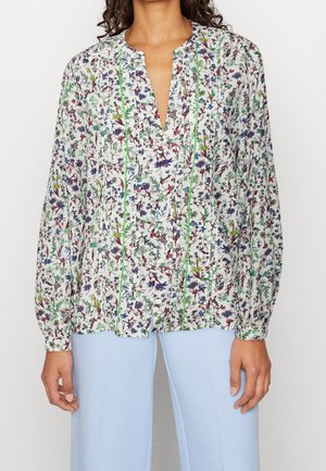 Frau trägt eine weiße Bluse mit langen Ärmeln, bedruckt mit bunten Blumen und grünen vertikalen Nähten, kombiniert mit hellblauen Hosen.