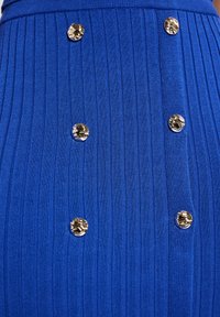 Tissu côtelé bleu royal avec des boutons décoratifs en or, présentant des lignes verticales et une coupe ajustée. Texture lisse avec des accents brillants.