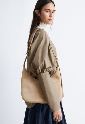 Kvinde med langt mørkt hår iført beige bluse med pufærmer, hvid rullekrave, marineblå nederdel, bærende på stor beige skuldertaske, kigger til siden.