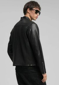 Zwarte leren jas met een nauwsluitend ontwerp, voorzien van een hoge kraag, ritsen aan de manchetten en minimalistische hardware accenten.