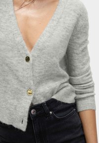 Cardigan grigio chiaro realizzato in materiale morbido e peloso. Presenta un profondo scollo a V, bottoni dorati e maniche aderenti, abbinato a jeans in denim scuro.